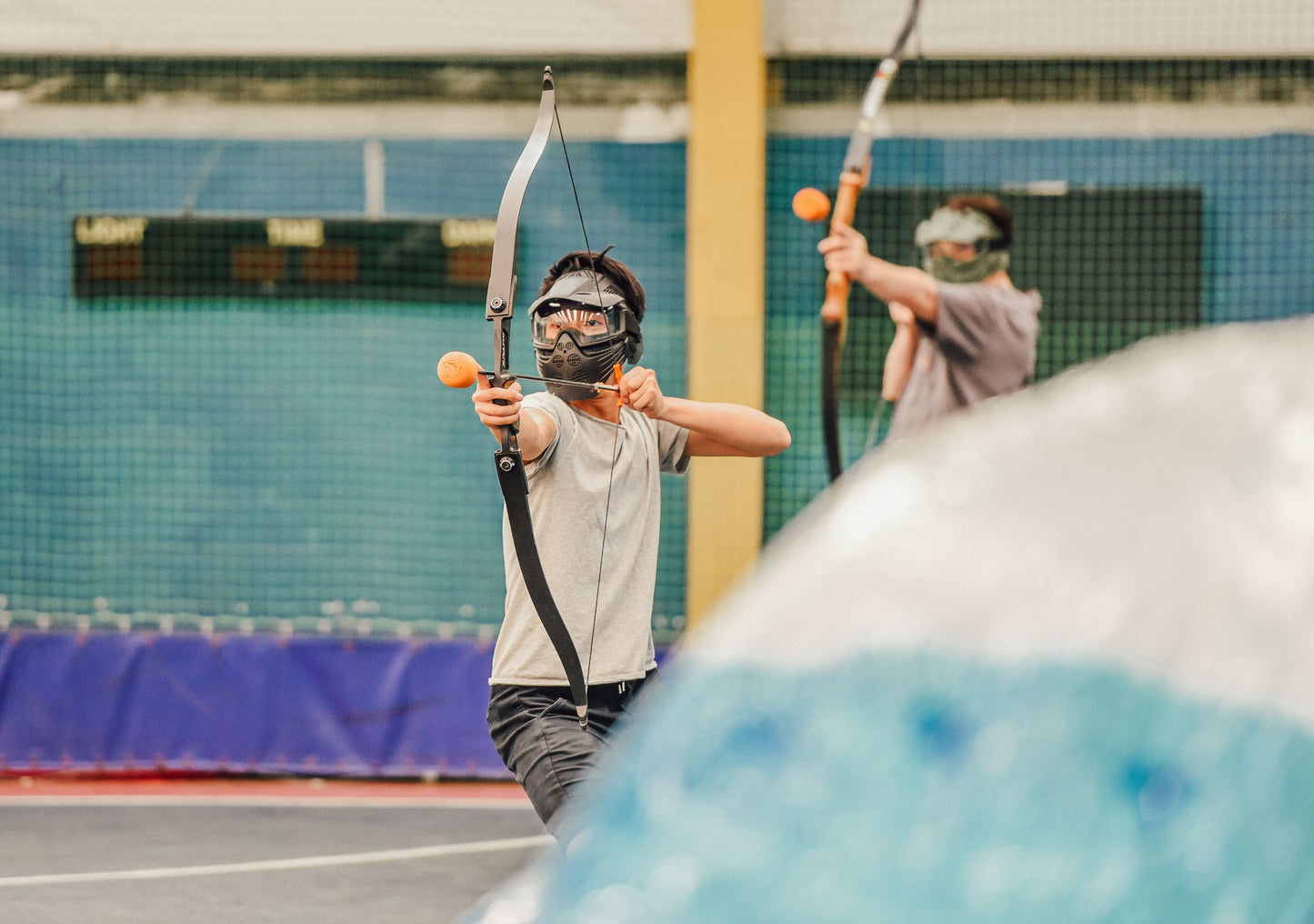 Two people practicing archery in an indoor setting with protective gear.