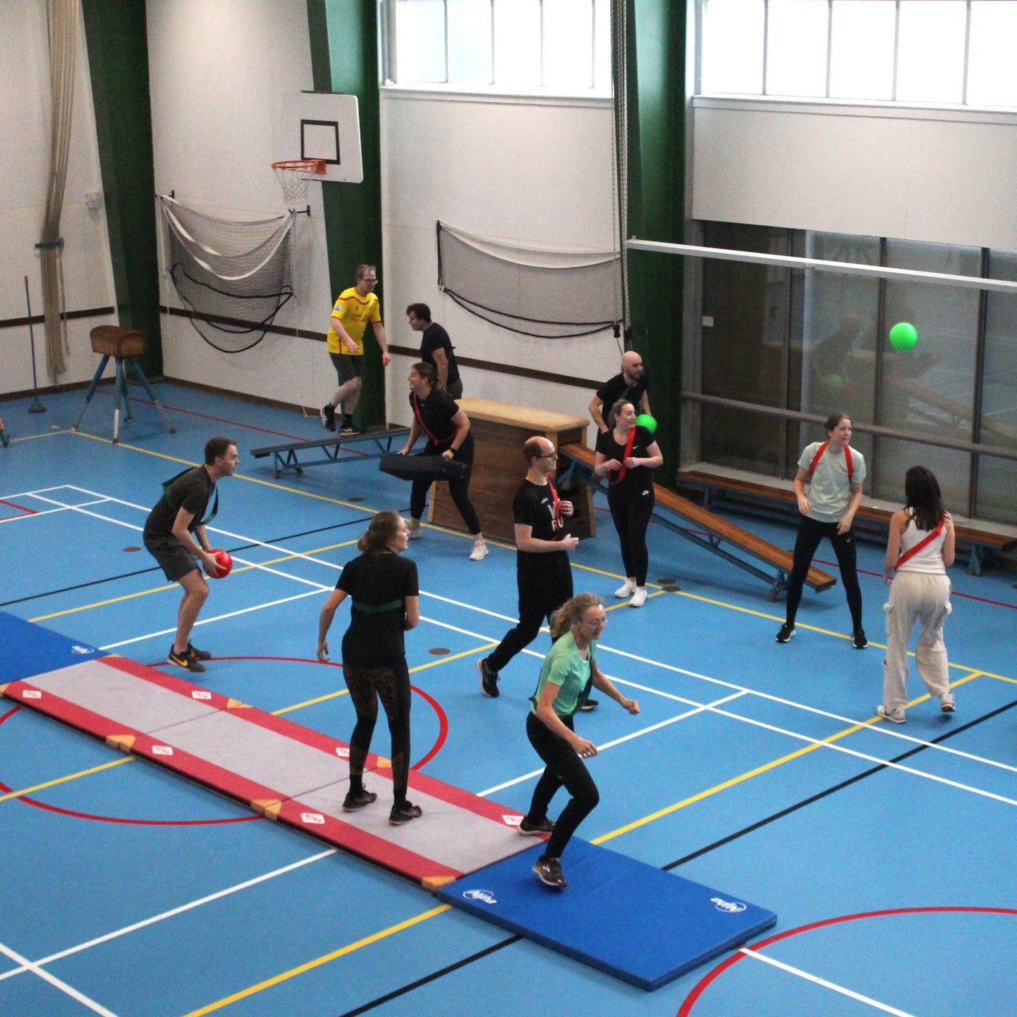 Group of people playing dodgeball in a gymnasium with sports equipment around.
