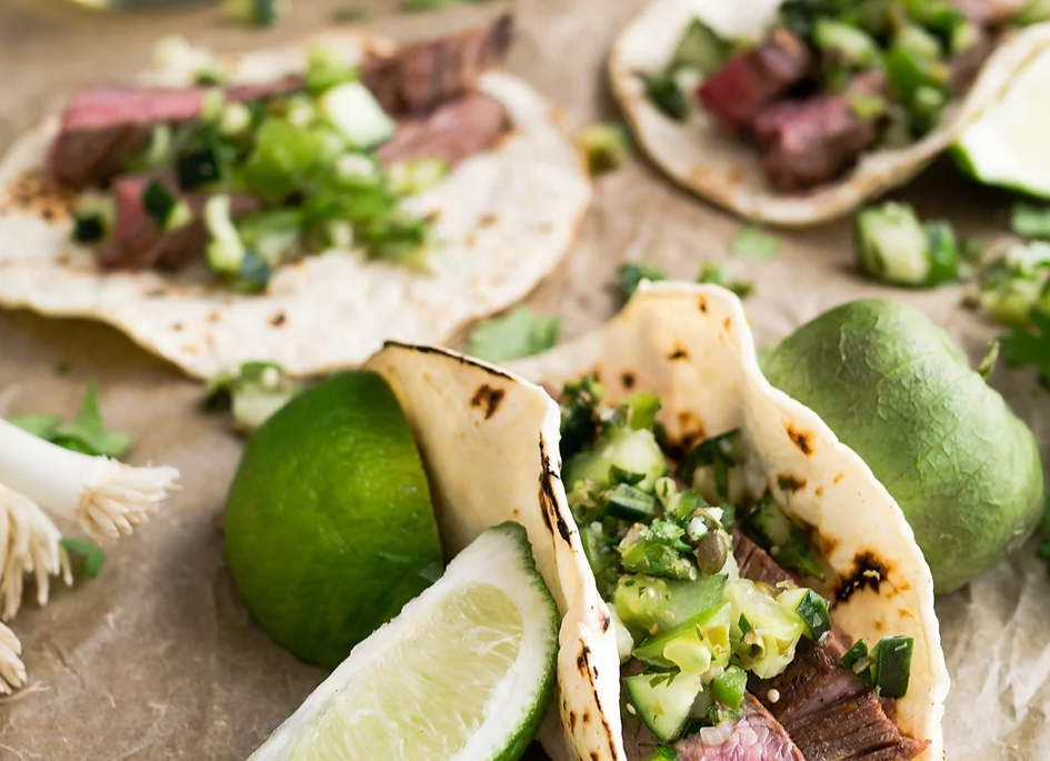 Tacos with meat and vegetables on a wooden surface with limes.