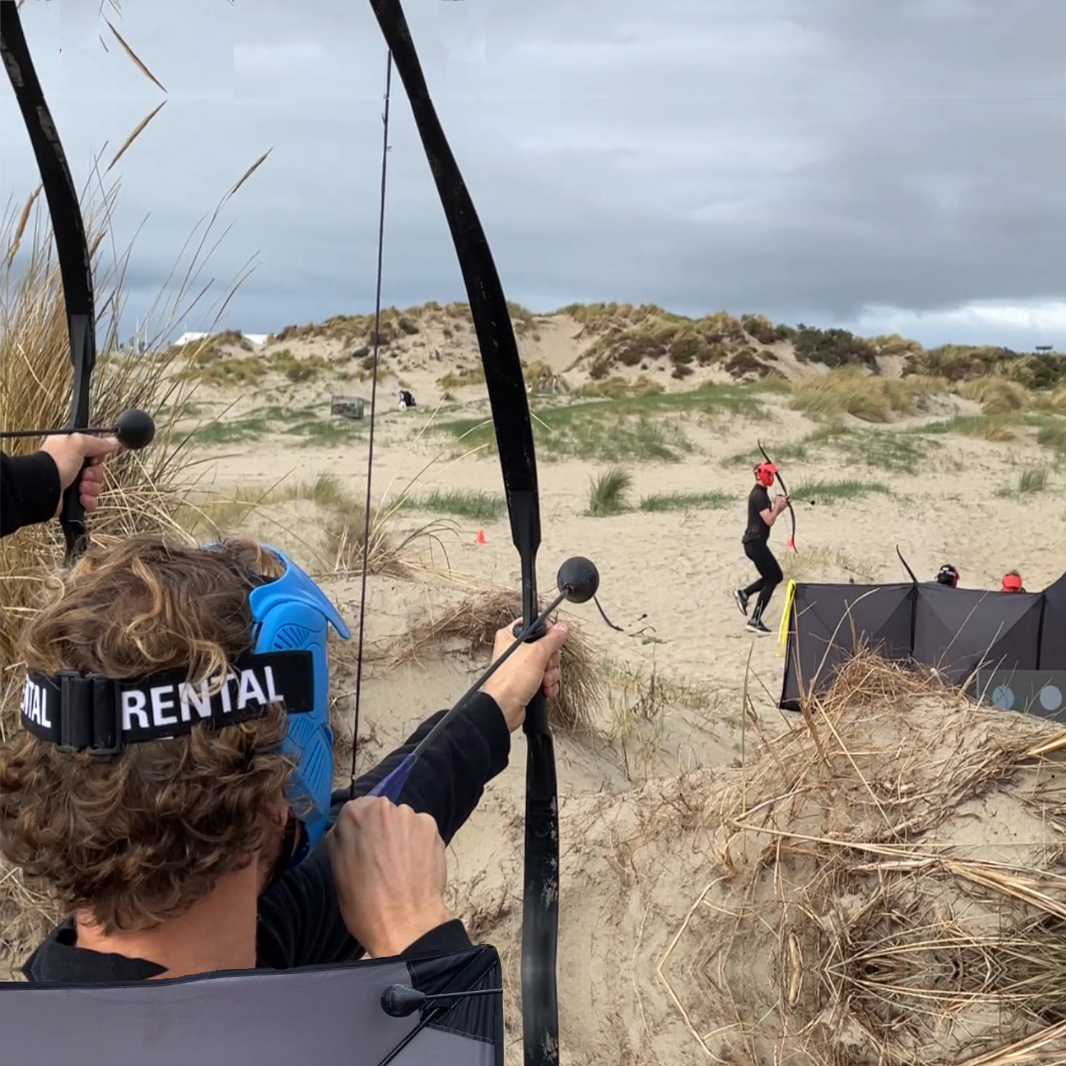 Person holding a bow and arrow on a sandy dune with other people in the background.